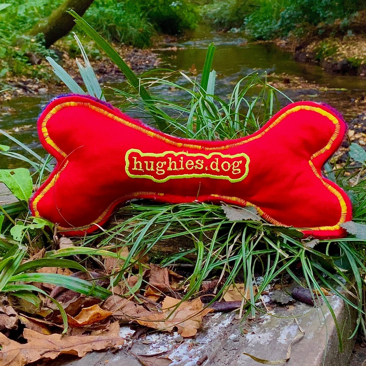 Red bone-shaped dog toy with Hughies Dog Accessories branding on grass near a stream.