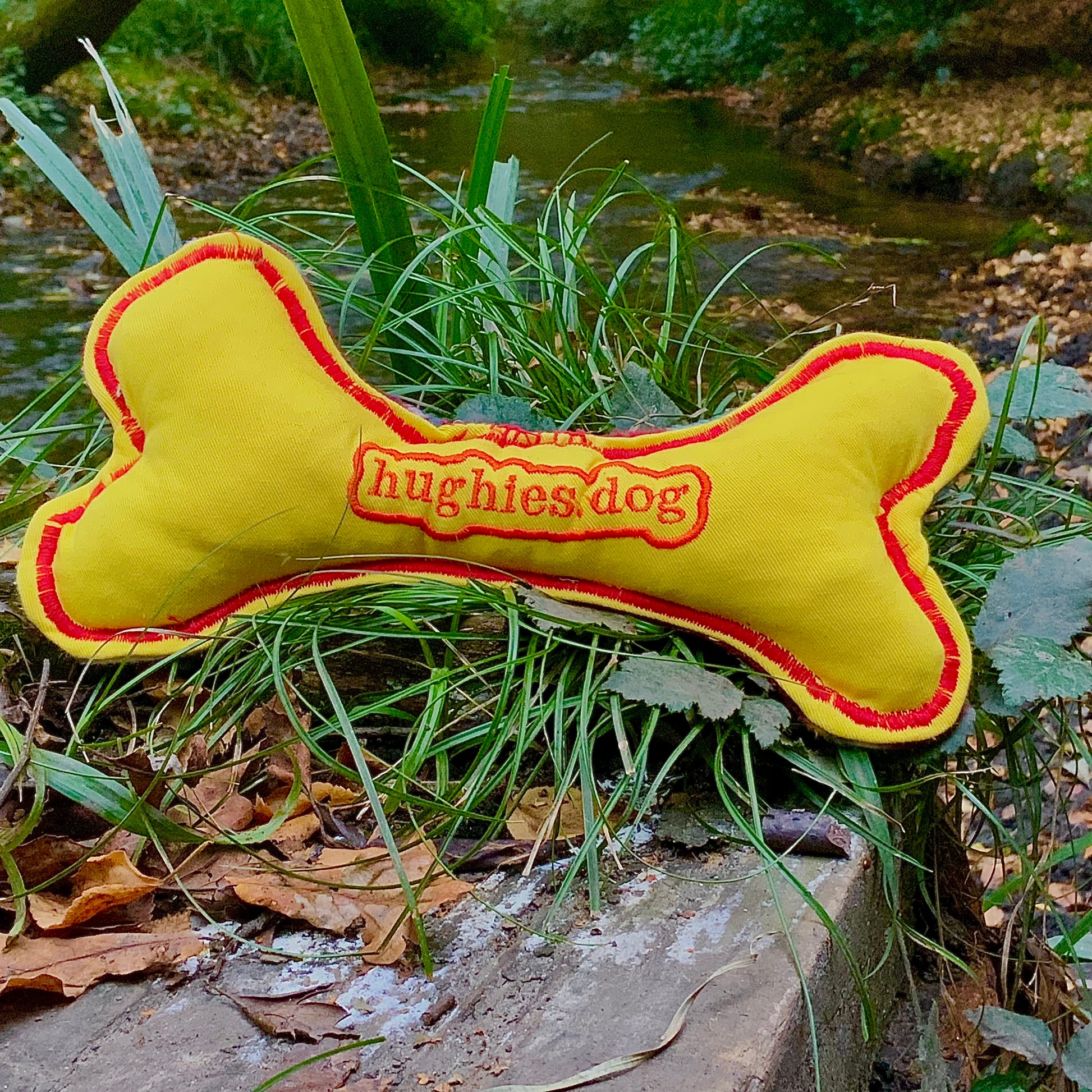 Yellow bone-shaped dog toy with red border and 'hughies.dog' branding on a natural background.